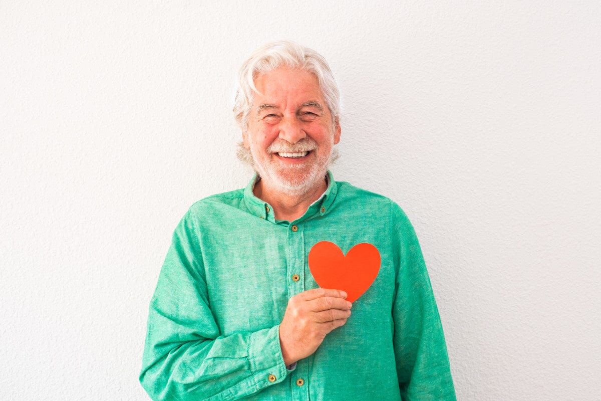 senior man smiling and holding a red paper heart.
