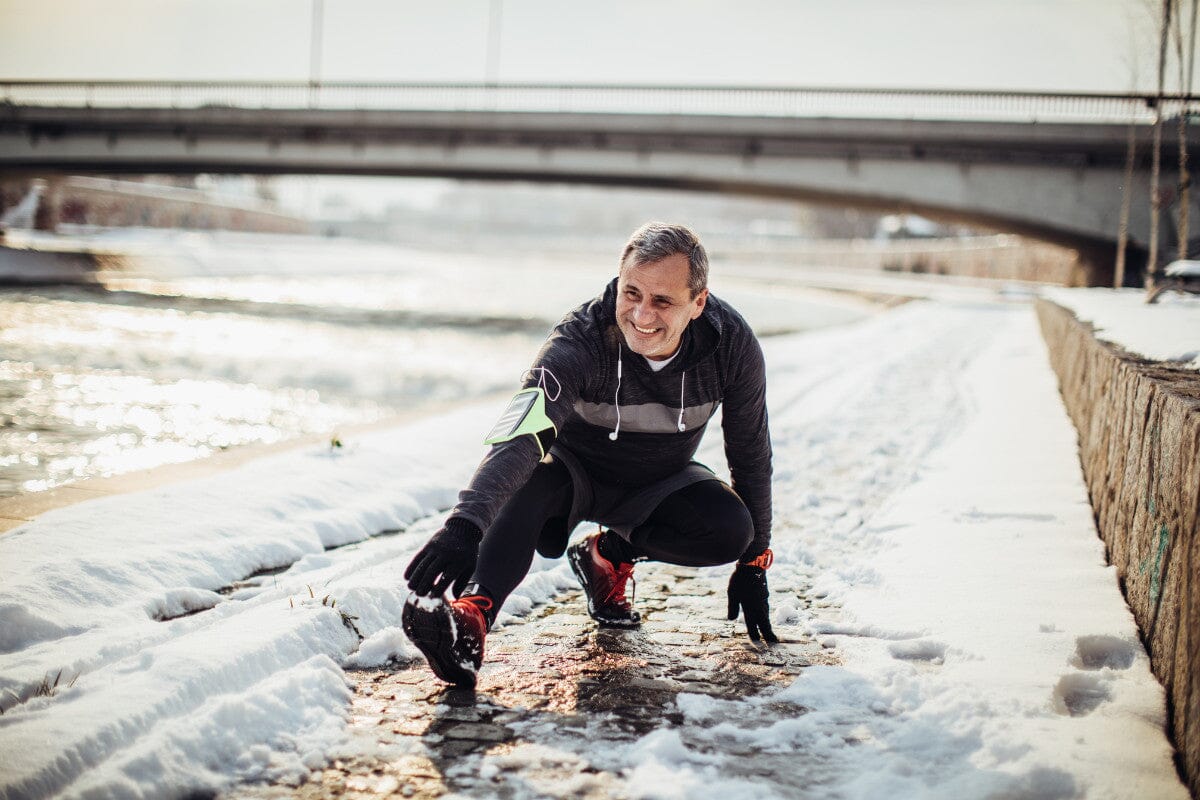 senior man stretching his leg in the snow alongside a river.