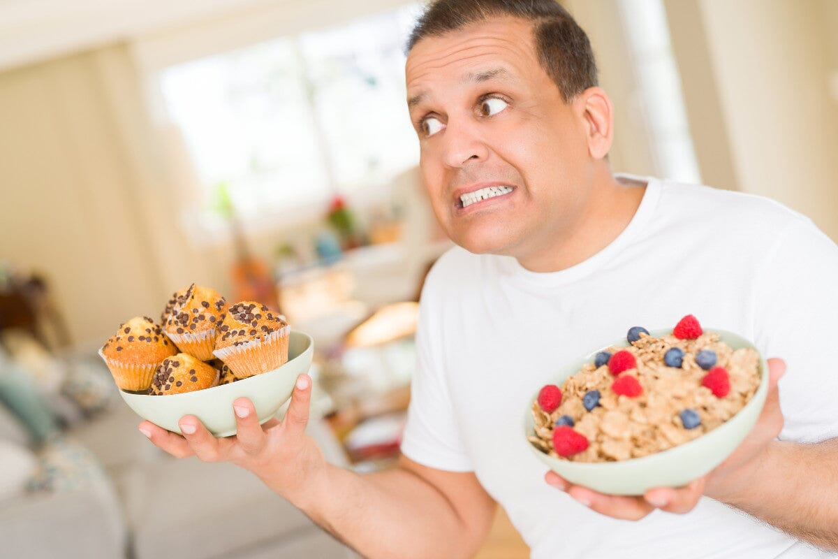 older man standing in his kitchen, trying to choose between a bowl of healthy granola with fruit and a bowl of chocolate chip muffins