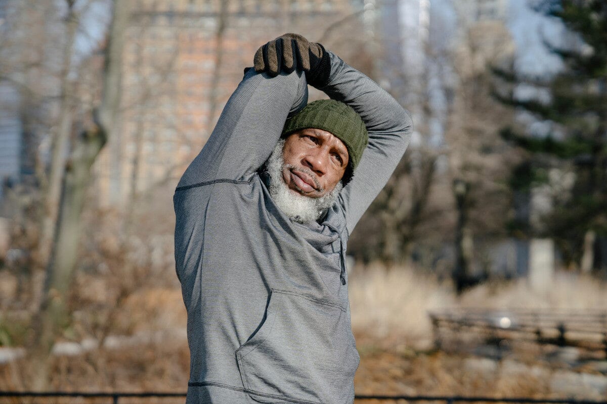 senior man dressed in a gray sweater and green beanie, stretching outside in the cold weather.