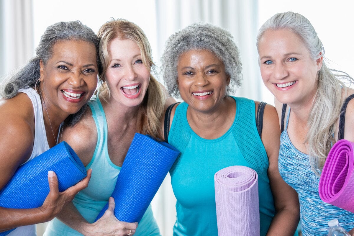 4 older women wearing active clothing and holding yoga mats, smiling at the camera