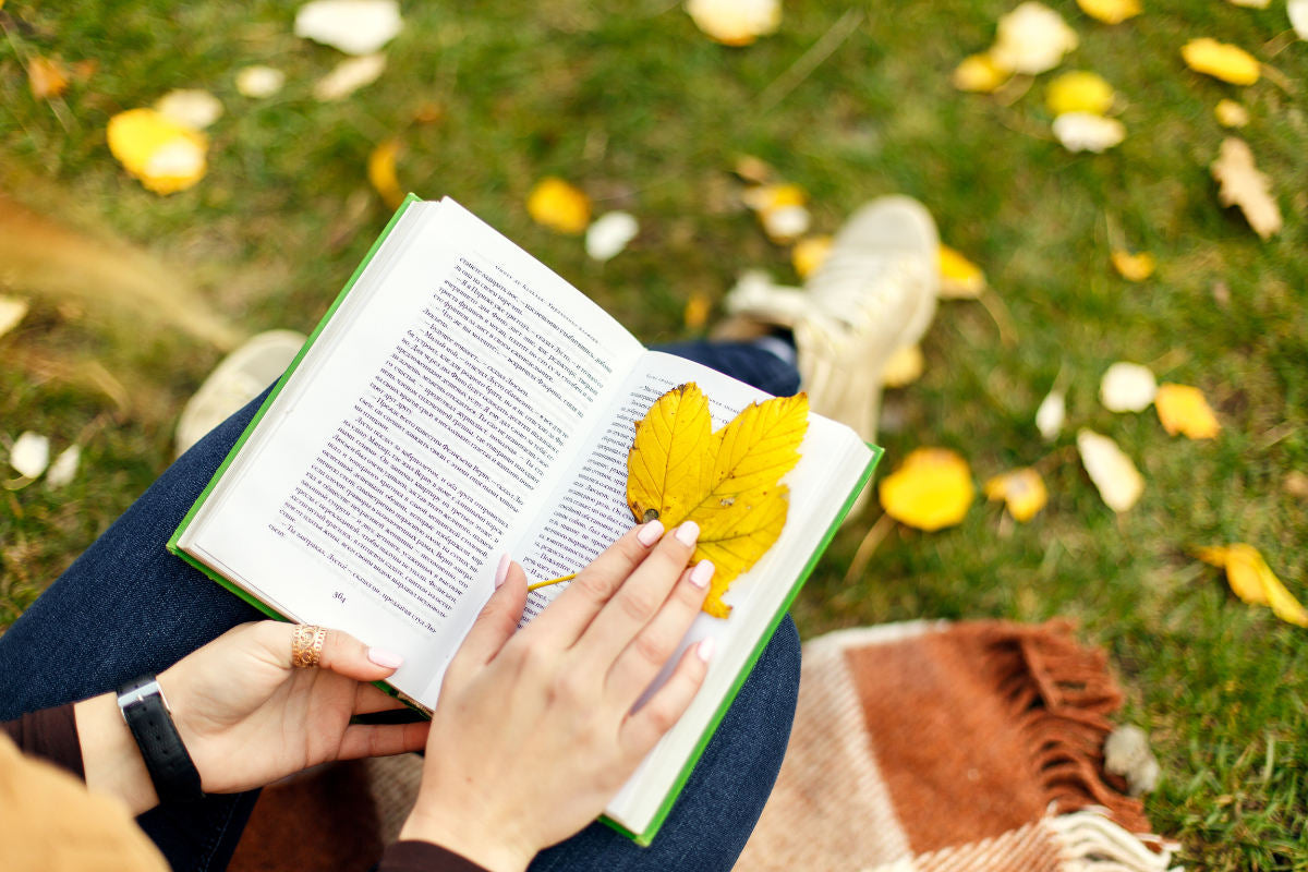 woman's hands on open book. yellow leaf sitting on right page of book. background is green grass with yellow leaves on it.
