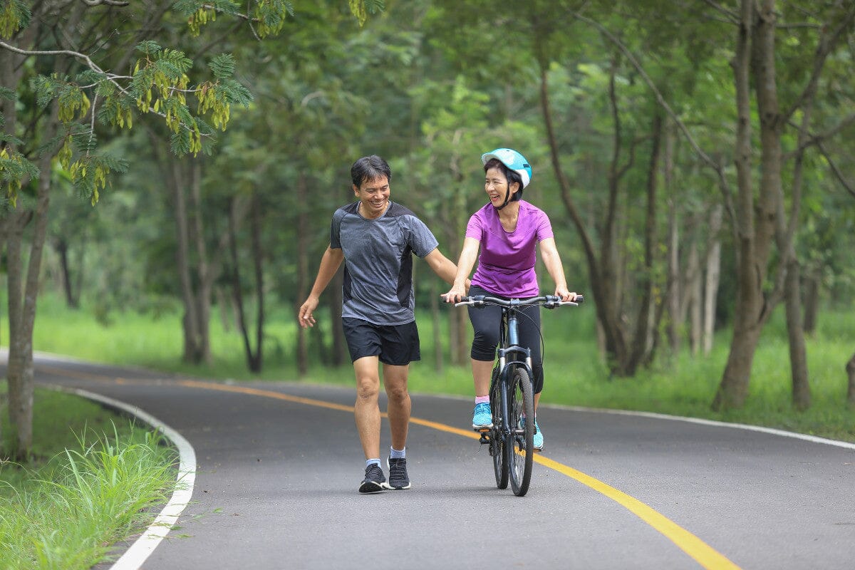 woman riding a bike next to a man running on the road surrounded by trees