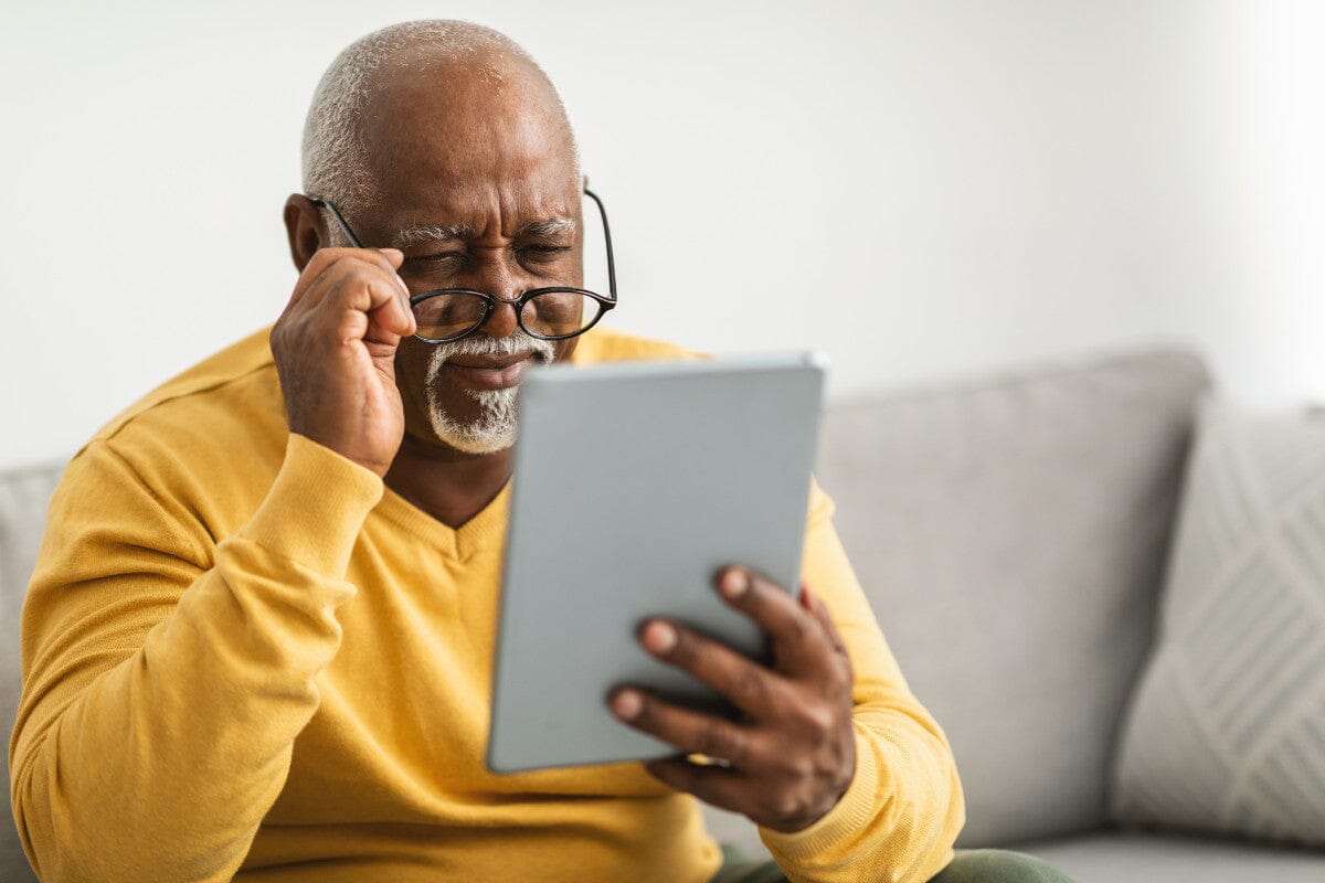 older man sitting on the couch, holding his glasses as he squints to read on his iPad