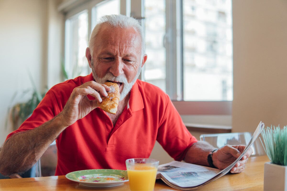 Senior man in a red collar shirt, reading the news while eating a pastry for breakfast.