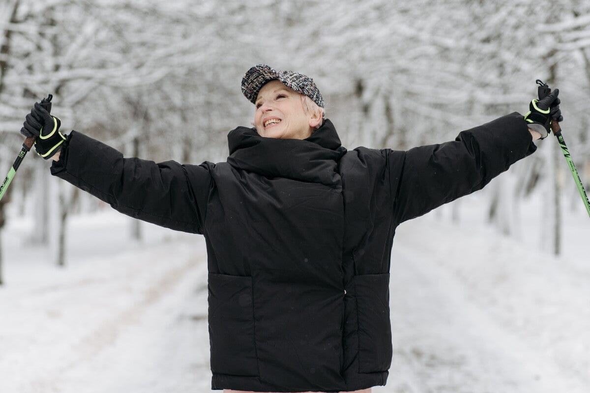 senior woman wearing a black hat and big black coat, holding walking sticks while she stands outside in the winter snow