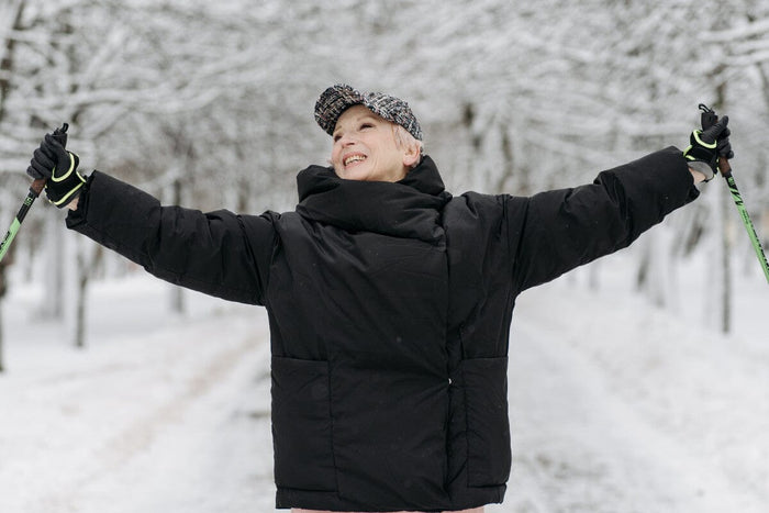 senior woman wearing a black hat and big black coat, holding walking sticks while she stands outside in the winter snow
