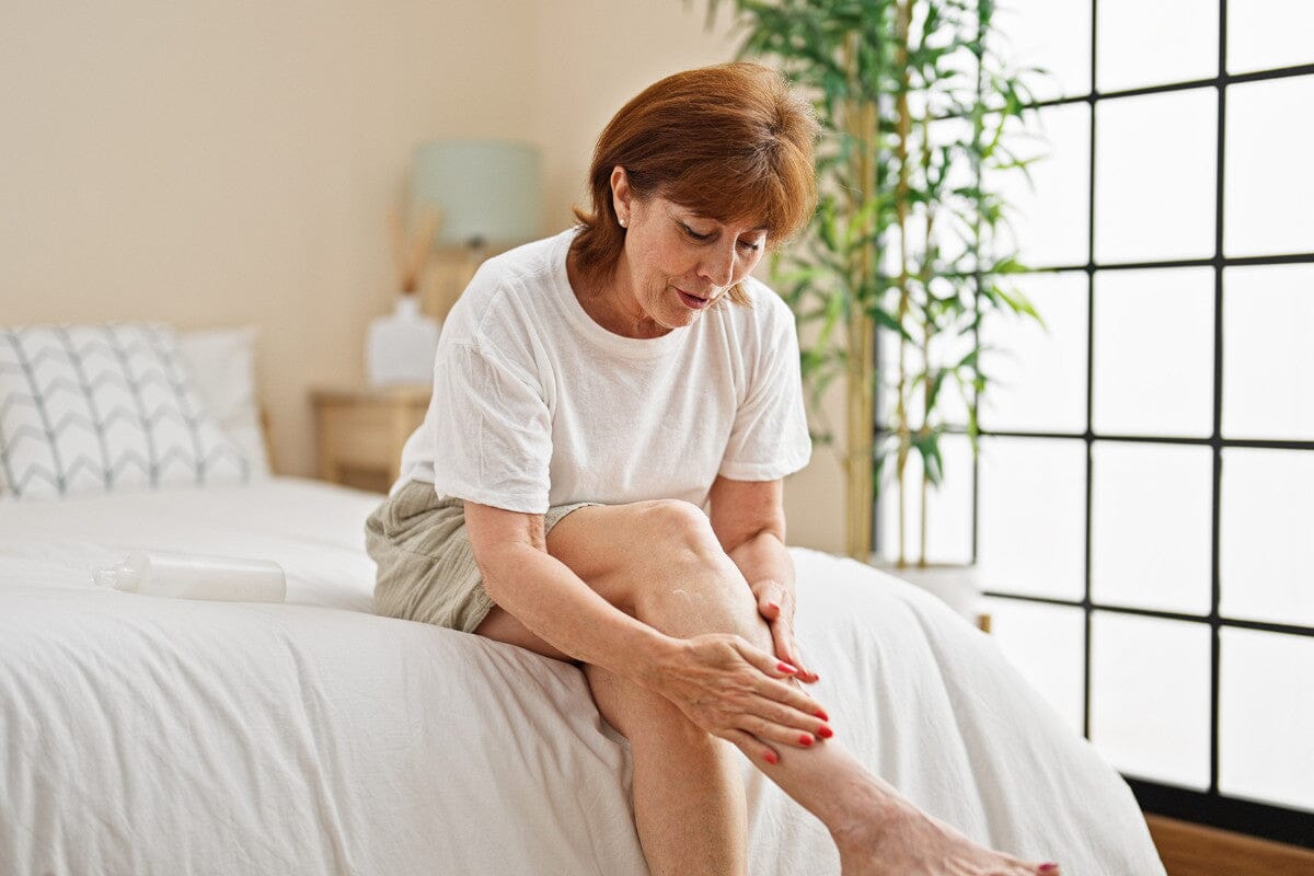 Middle age woman applying lotion on leg sitting on bed at bedroom