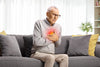 elderly man sitting on a gray couch in pain because his heart is inflamed.