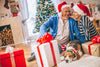 senior couple wearing santa hats sitting on the floor next to presents, a Christmas tree, and their sleepy dog.