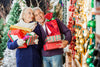 senior couple dressed in warm clothes, smiling and carrying wrapped presents as they look at the Christmas decorations outside.