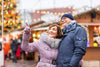 older couple wearing big coats and sightseeing in a town where there are Christmas decorations and a large Christmas tree