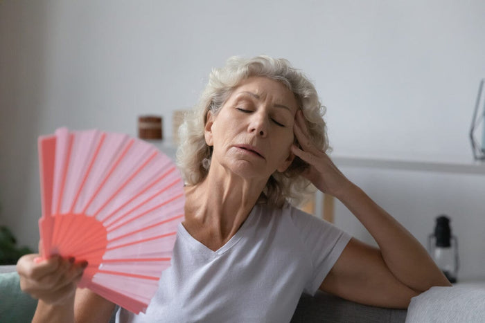 senior woman exhausted from the heat, cooling herself off inside with a handheld pink paper fan.