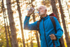 male senior taking a sip of water as he hikes through the woods in autumn.