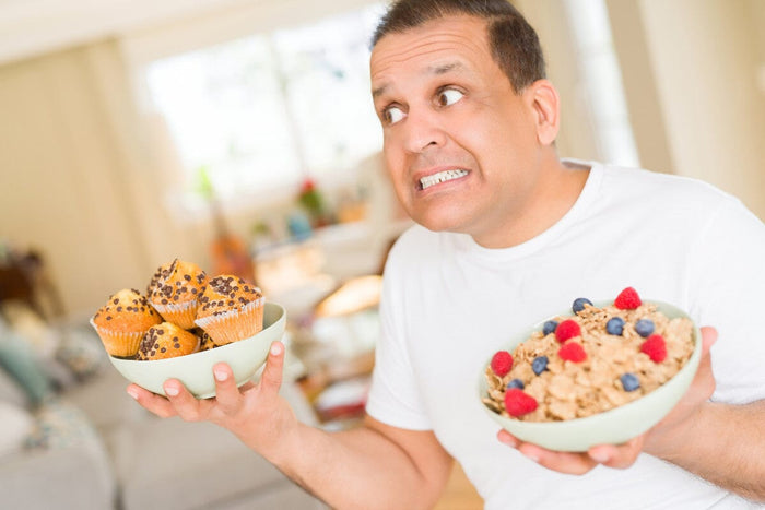 older man standing in his kitchen, trying to choose between a bowl of healthy granola with fruit and a bowl of chocolate chip muffins