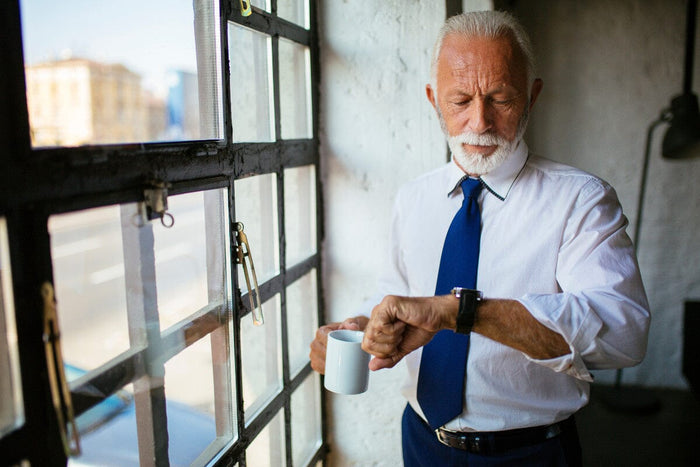 older man dressed in a suit and tie, standing next to a window in his apartment, holding a cup of coffee and looking down at his watch