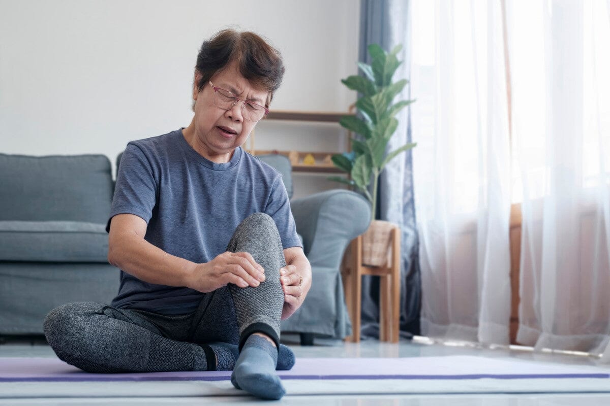 senior woman sitting on the living room floor, massaging her sore calf