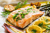 large piece of grilled salmon with green herbs laid next to slices of lemons.