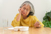 older woman wearing a yellow sweater staring down at a plate of food, looking like she's bored and uninterested in eating