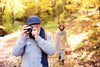 senior couple walking along a trail during the fall. senior man in front, holding up a camera, about to take a picture.