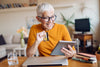 A senior woman in a yellow rolled-up long-sleeve shirt sits at a wooden desk with a closed laptop in front of her. She holds her iPad in one hand and her debit card in the other as she smiles gleefully because she just made a safe online holiday purchase!