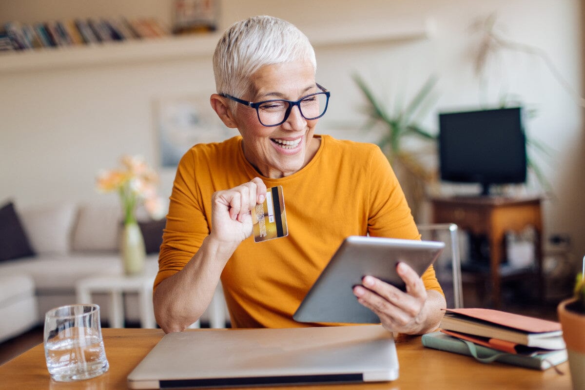 A senior woman in a yellow rolled-up long-sleeve shirt sits at a wooden desk with a closed laptop in front of her. She holds her iPad in one hand and her debit card in the other as she smiles gleefully because she just made a safe online holiday purchase!