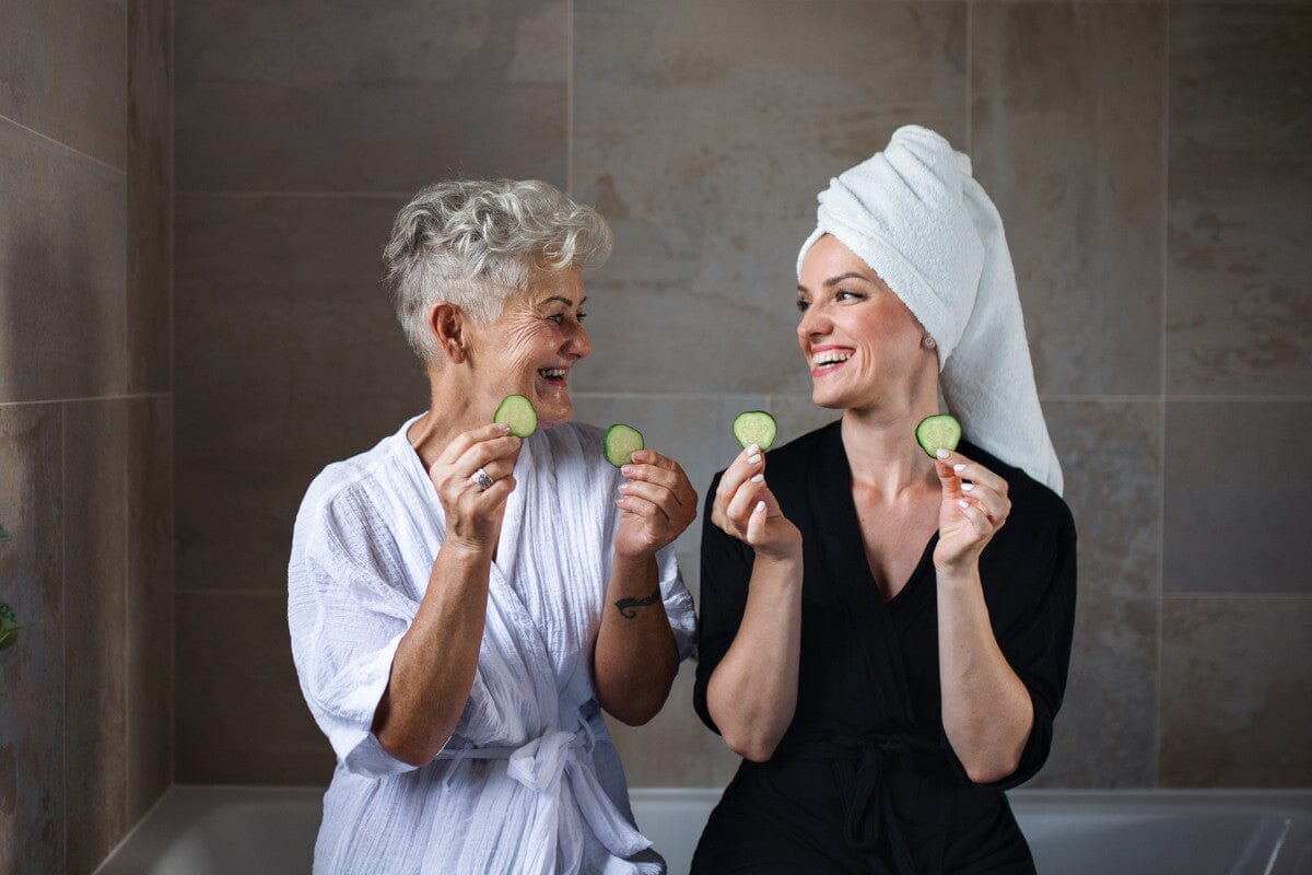 senior woman and mature woman sitting on the edge of a white bathtub holding up cucumber slices
