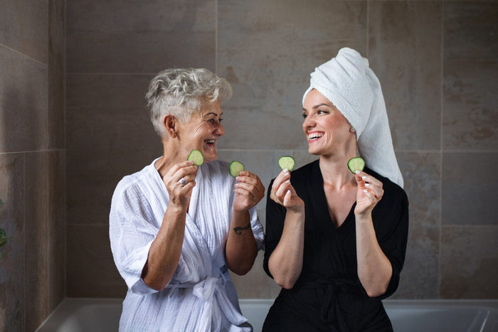 senior woman and mature woman sitting on the edge of a white bathtub holding up cucumber slices