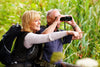 senior couple crouching in tall grass, as the woman points out something and the man looks through his binoculars.
