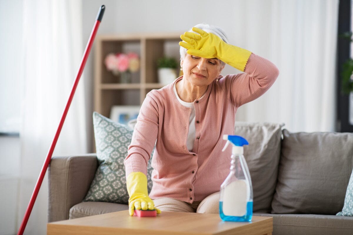 Senior woman tired from spring cleaning, sitting on the couch as she wipes down a table.