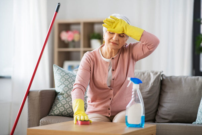 Senior woman tired from spring cleaning, sitting on the couch as she wipes down a table.