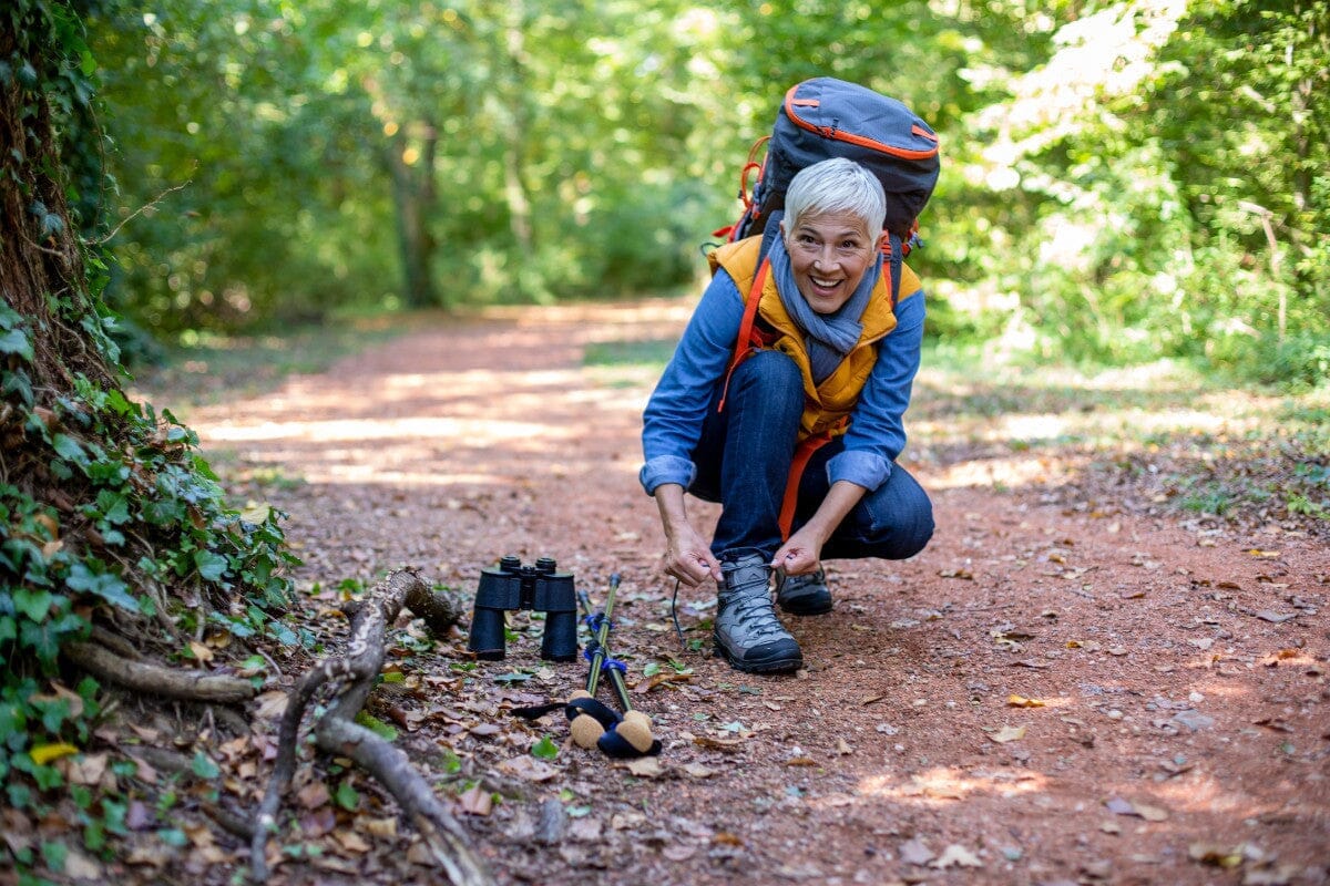 Senior woman on a hiking trip in spring, knelt down to tie her shoe on a leafy trail.