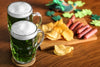 Two glasses of green beer and a board with chips and sausages on a wooden table.