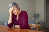 older woman leaning on a wooden table, holding her hand to her head as if she's stressed.