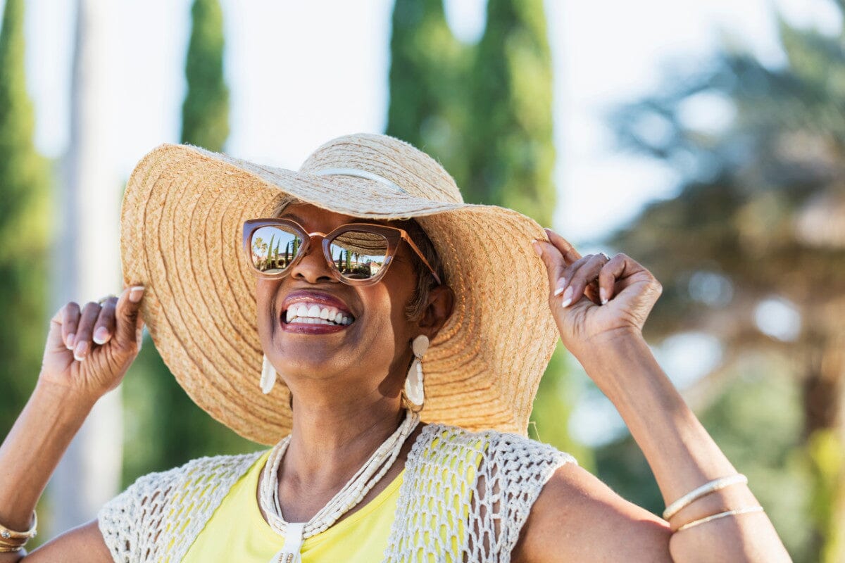 senior woman smiling and wearing a sun hat and sunglasses outdoors.