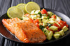 cooked salmon with avocado tomato salsa and sliced lemons placed neatly on a black dinner plate.