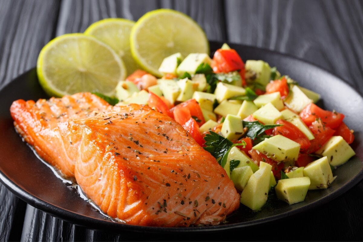 cooked salmon with avocado tomato salsa and sliced lemons placed neatly on a black dinner plate.