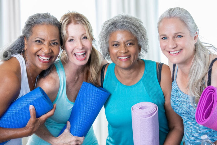 4 older women wearing active clothing and holding yoga mats, smiling at the camera