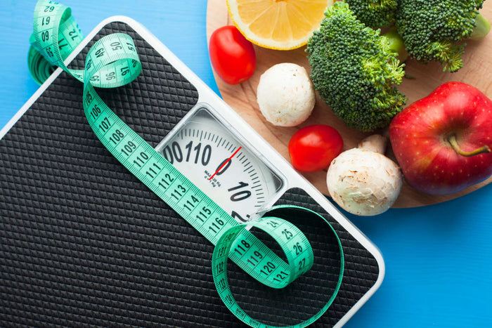 bodyweight scale and tape measure sitting next to a plate of fruits and vegetables on bright blue background