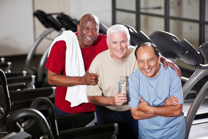 3 older men wearing workout clothes and standing in front of treadmills, smiling at the camera